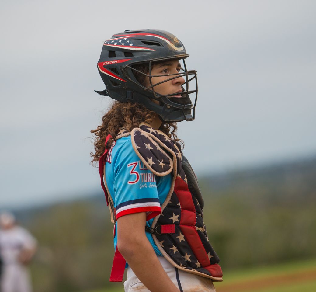 A softball player in a red and blue uniform stands ready at their position wearing protective catching gear.