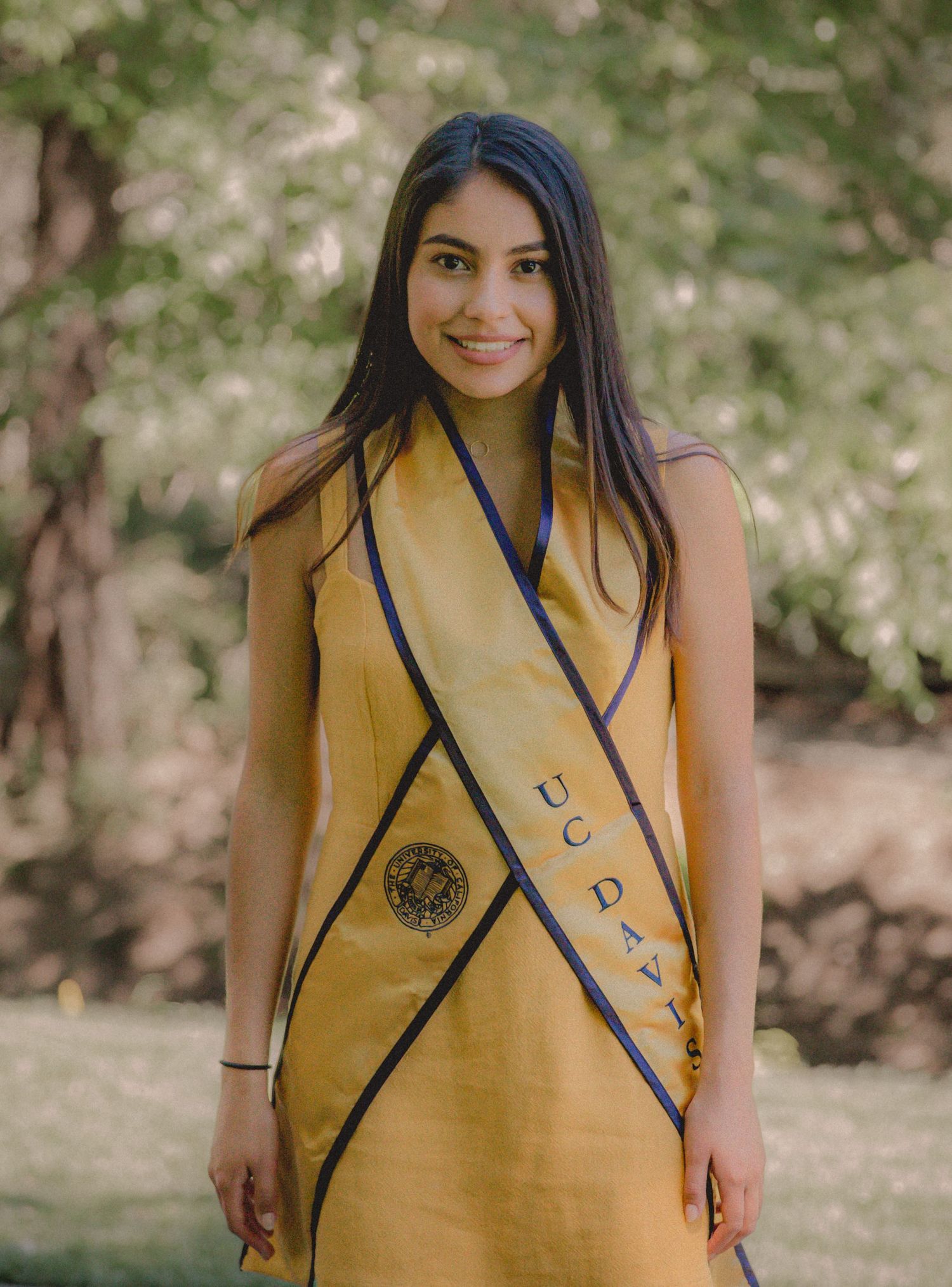 Graduate wearing gold academic stole with blue trim poses in outdoor setting.