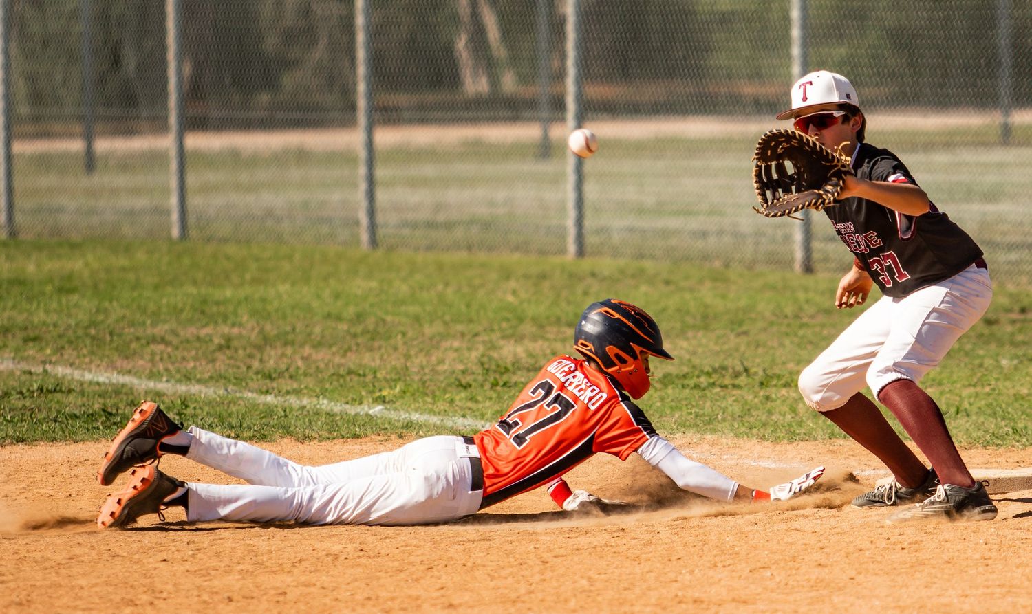Baseball player slides into base while fielder attempts to make the tag during competitive game on dirt infield.