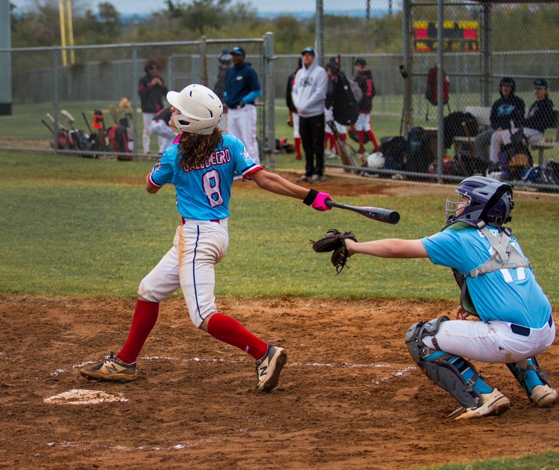 Youth baseball player in light blue jersey swings at pitch while catcher crouches during game on dirt diamond.