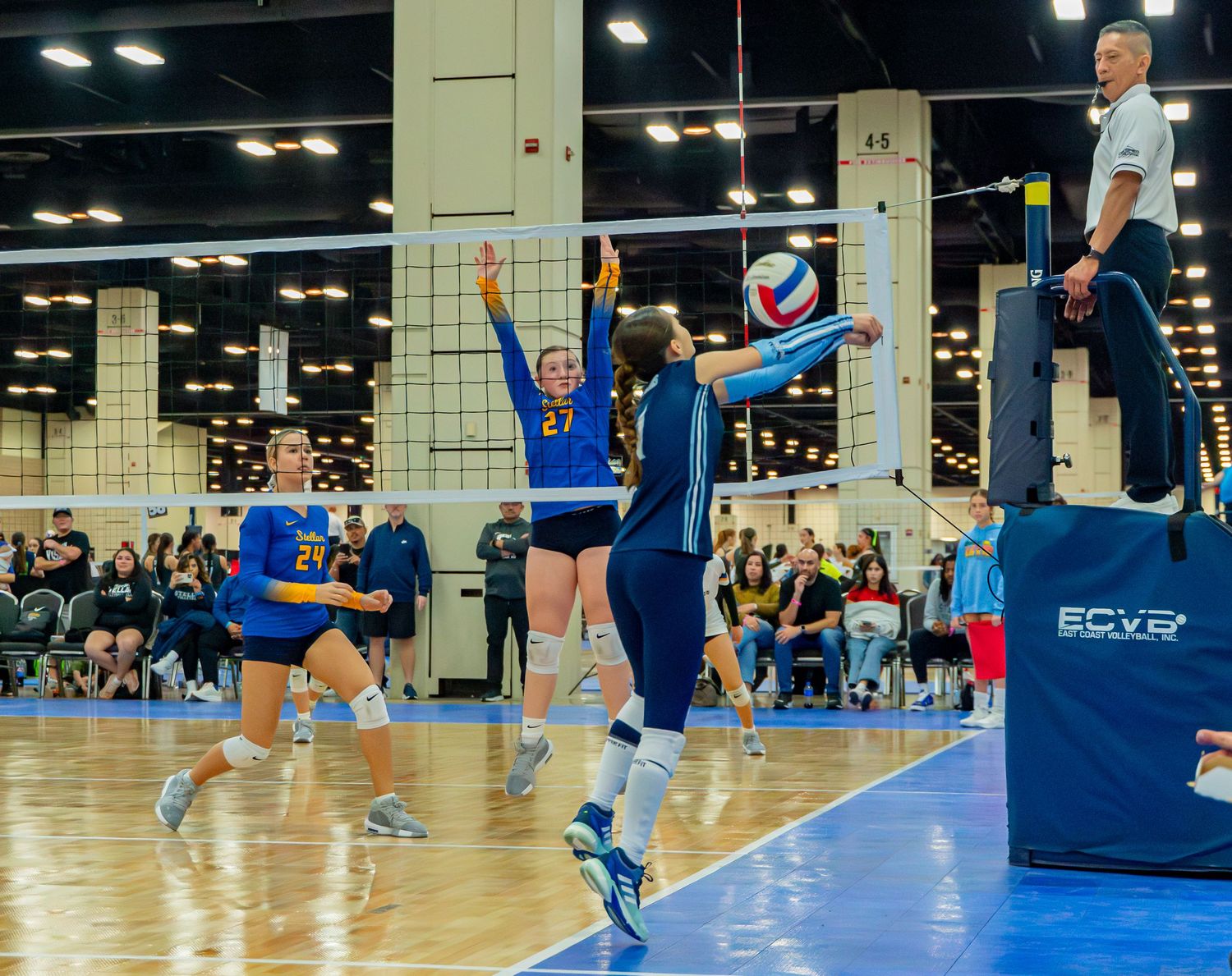 Indoor volleyball match showing players in blue uniforms jumping and diving for the ball.