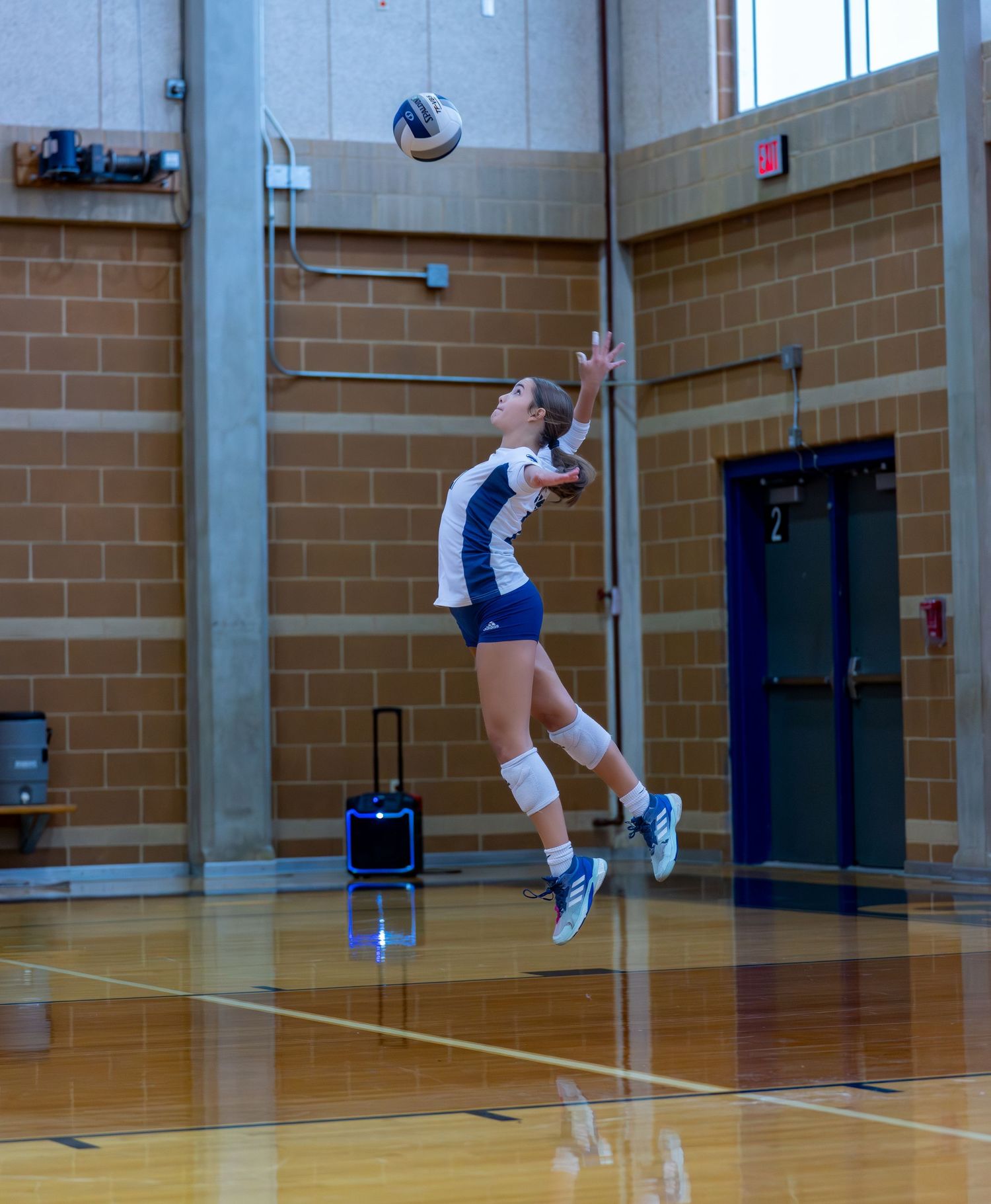 Player leaps high to serve volleyball in indoor gymnasium setting.