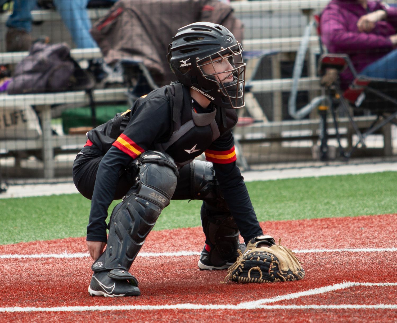Baseball catcher in dark protective gear crouching behind home plate during game.