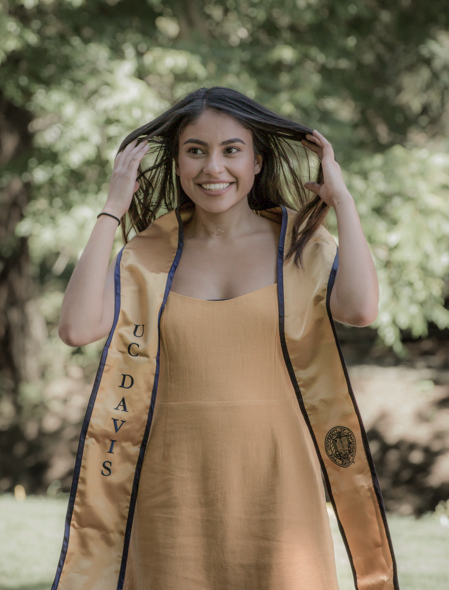 Graduate wearing yellow dress and blue graduation stole poses outdoors in natural setting.