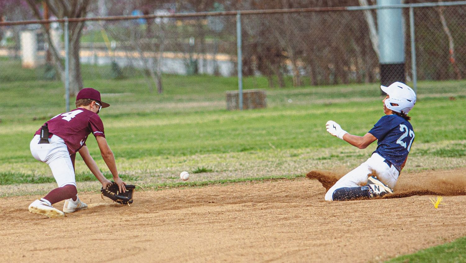Baseball fielder reaches for ground ball as runner slides into base.