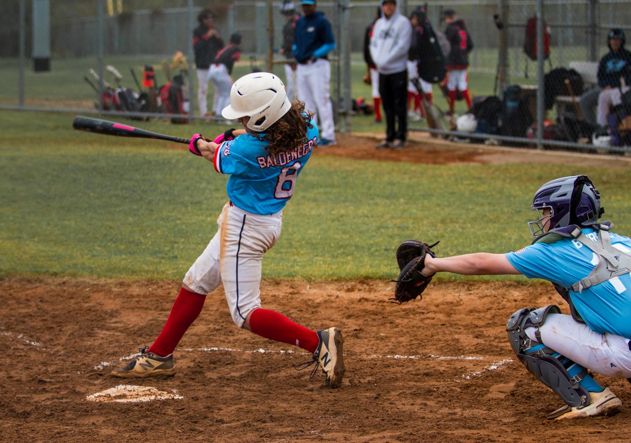 Youth baseball player in blue jersey and red socks swings at pitch while catcher awaits in defensive position.