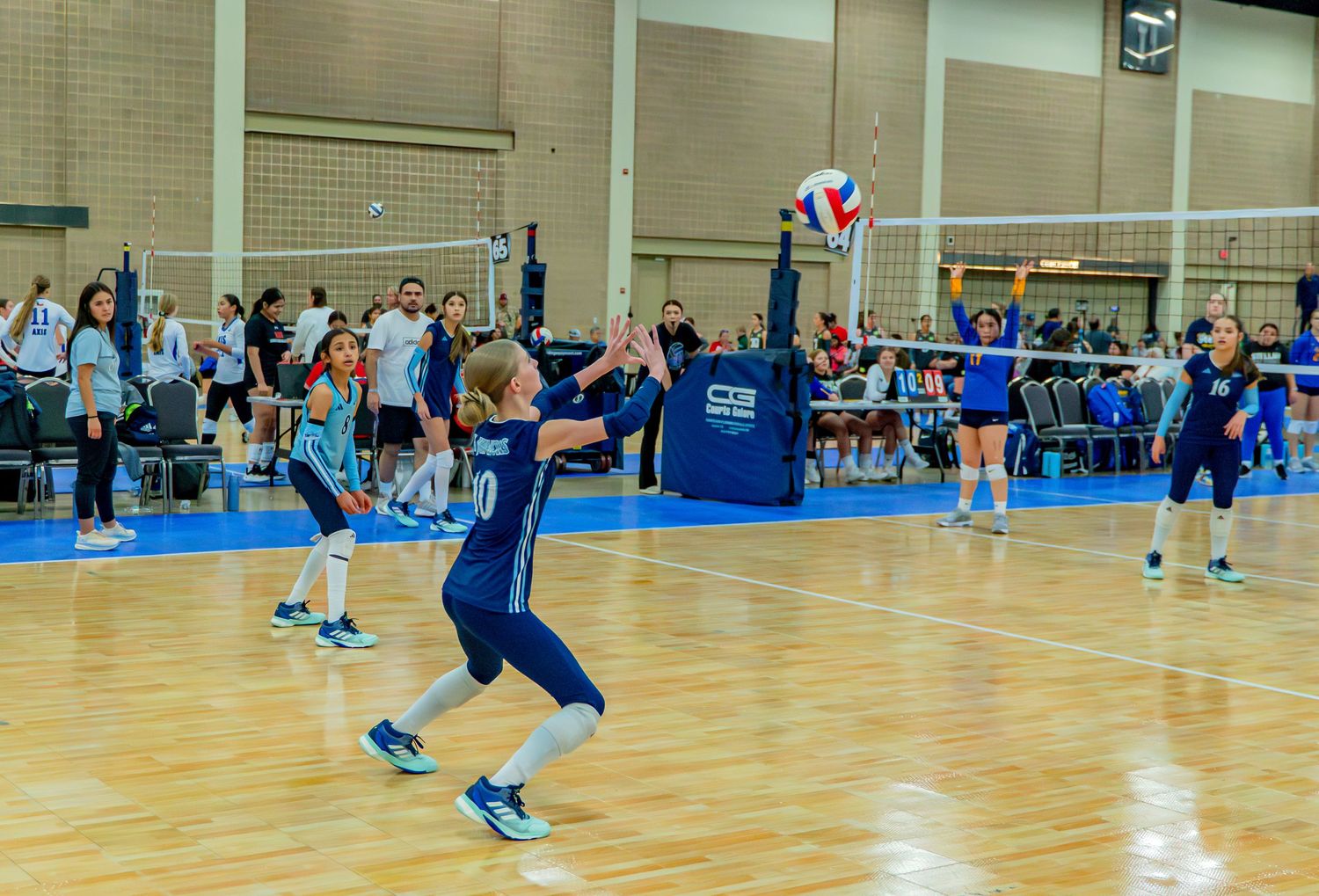 Indoor volleyball players in blue uniforms diving and jumping during an intense match on a bright blue court.