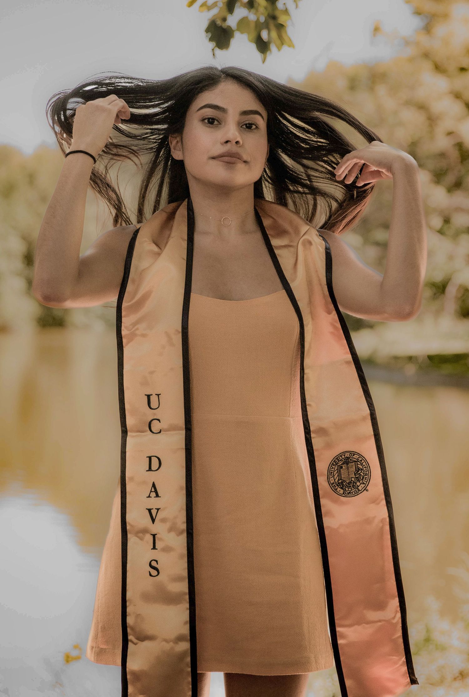 Graduate in tan dress with graduation stole poses in outdoor setting with trees and water in background.