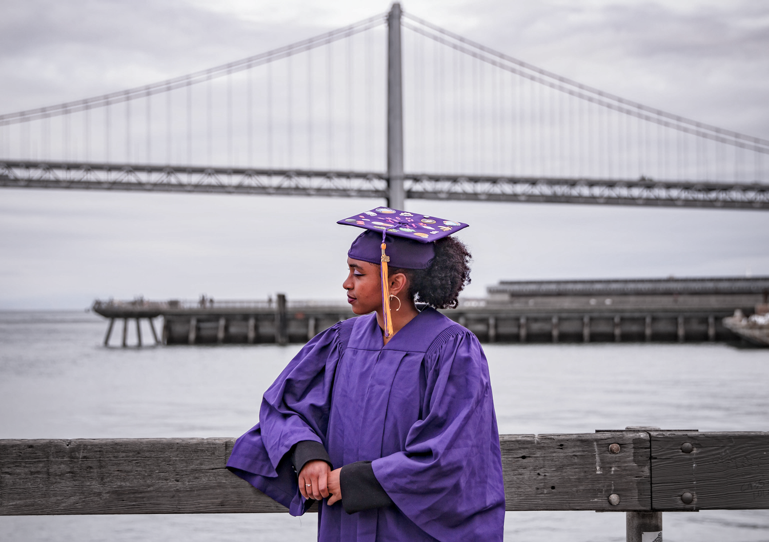 Graduate in purple graduation gown poses with San Francisco Bay Bridge in background.