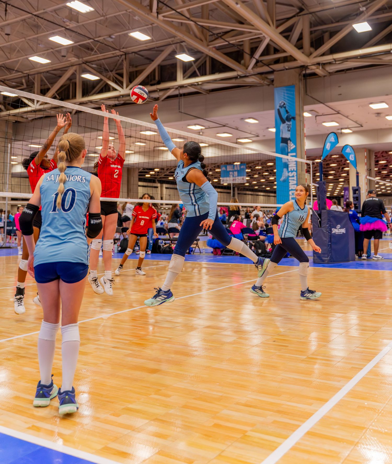 Wide view of a volleyball match in progress on an indoor court with players in blue and red uniforms.