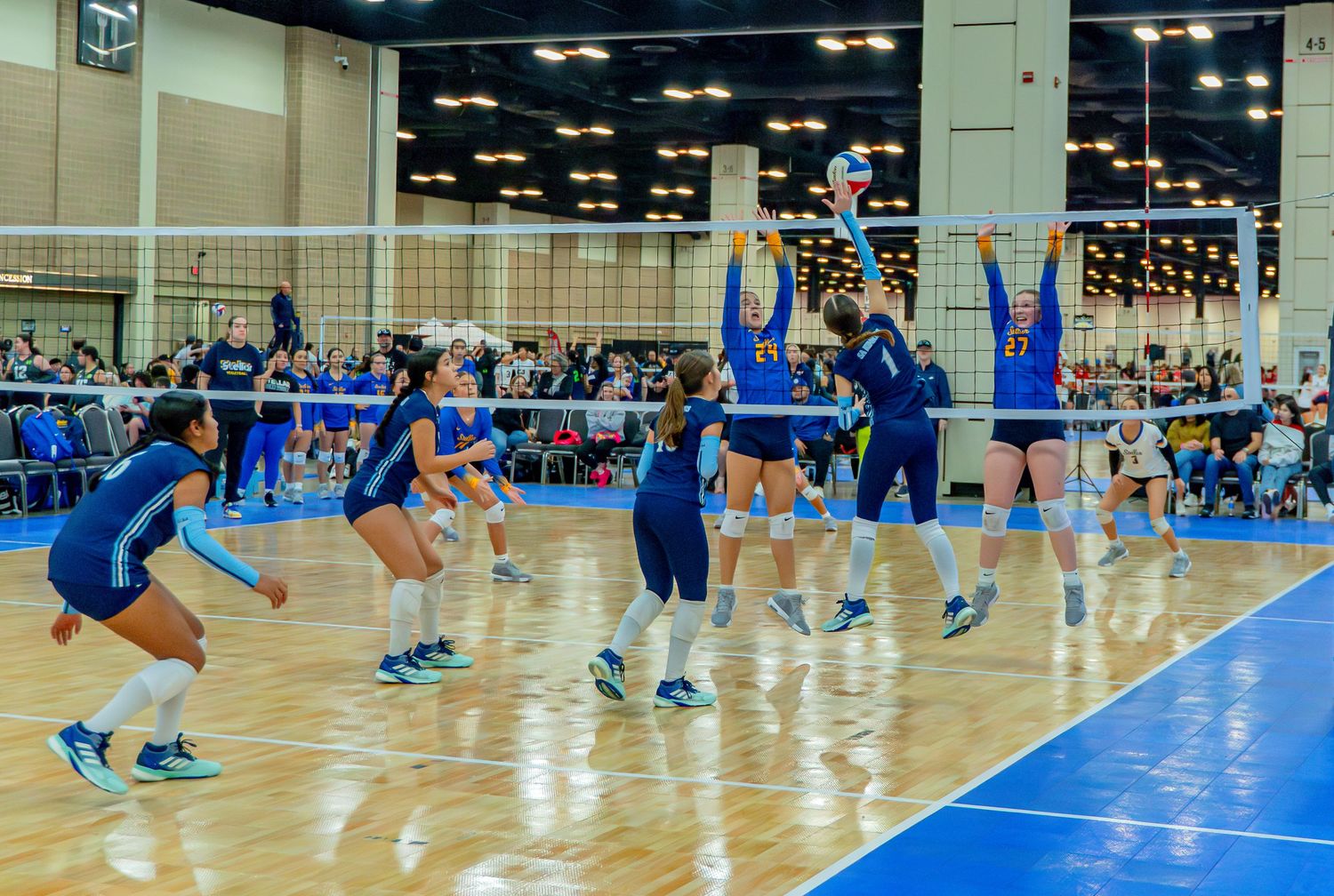 Wide panoramic view of volleyball players competing on indoor blue court with spectators watching.