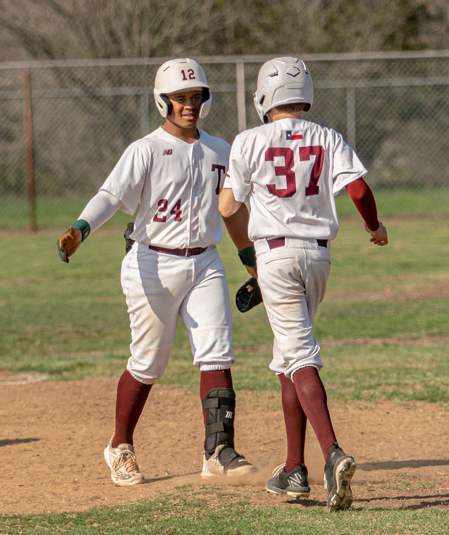 Youth baseball players in white uniforms running bases during daytime game.