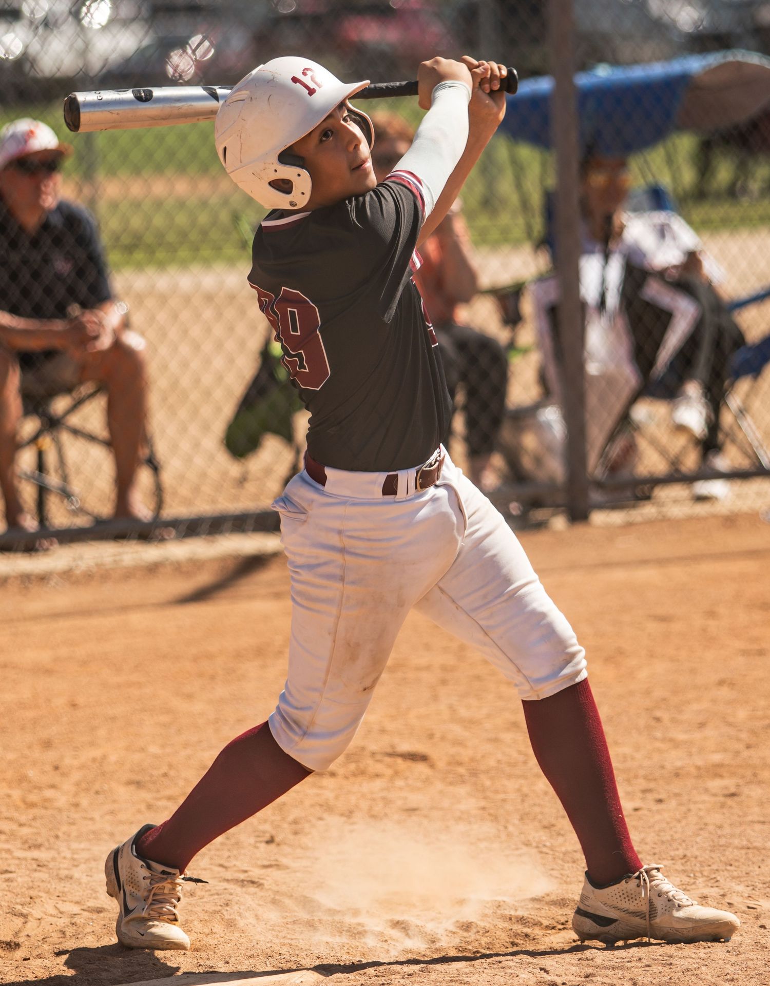 Baseball batter in dark uniform swings bat during game with spectators in background.