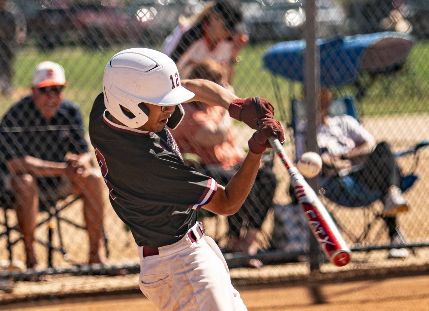 Baseball batter in white uniform swings bat during game with spectators visible behind fence.