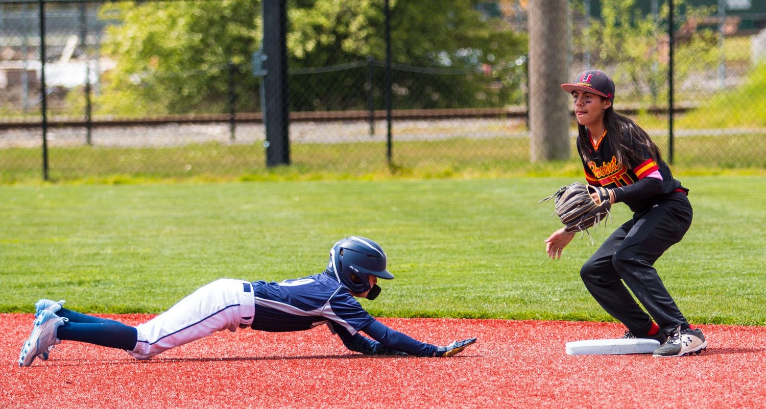 Baseball player in blue slides into base while defender in black uniform prepares to make the play.