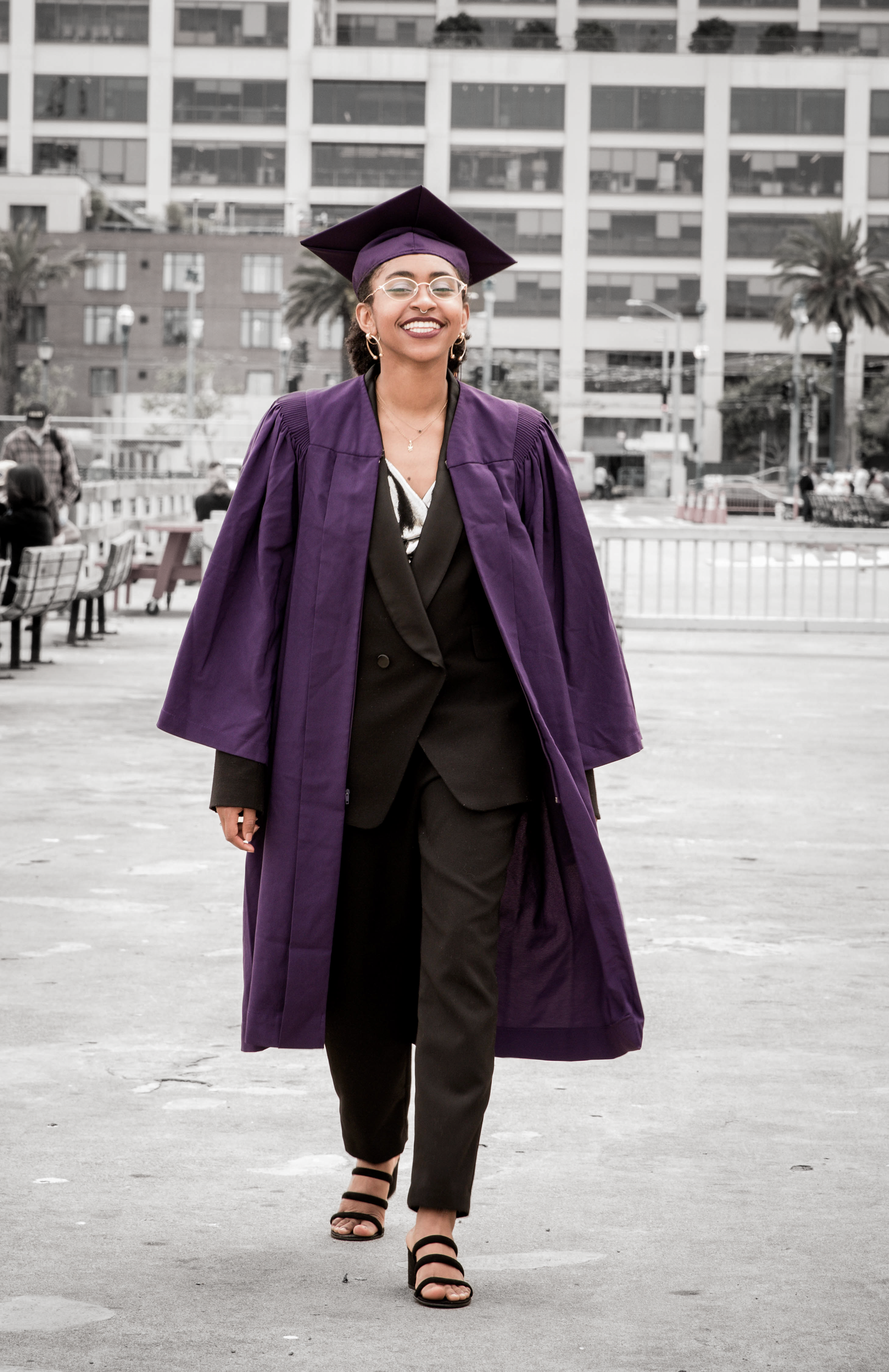 Graduate in purple cap and gown walking confidently across outdoor plaza.