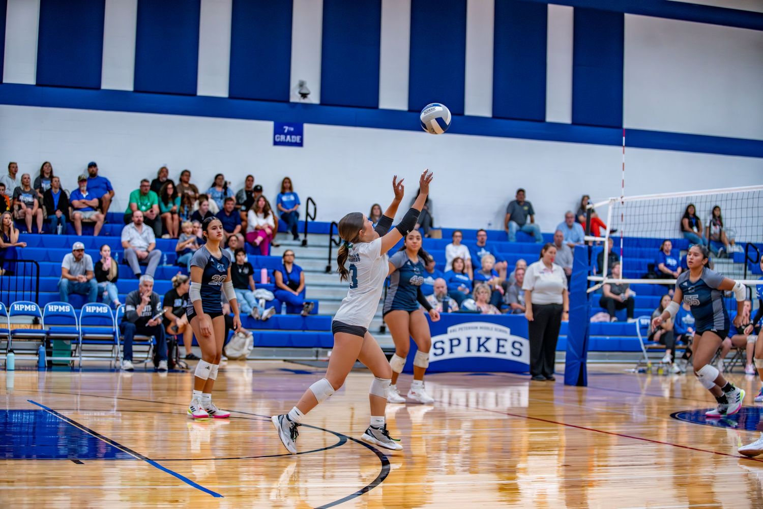 Wide view of volleyball match showing players and spectators in blue and white gymnasium.