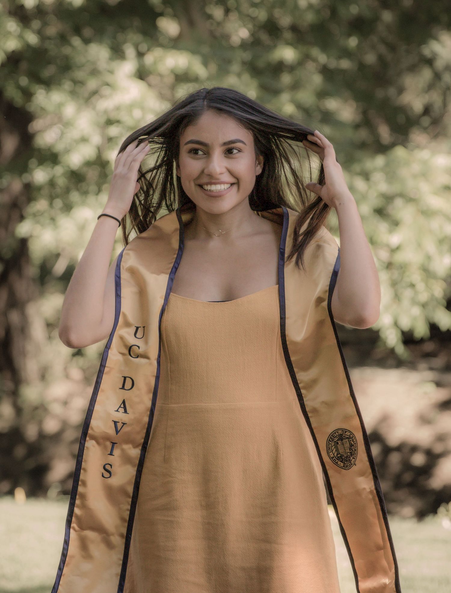 Graduate wearing yellow dress and blue graduation stole poses outdoors in a park setting.