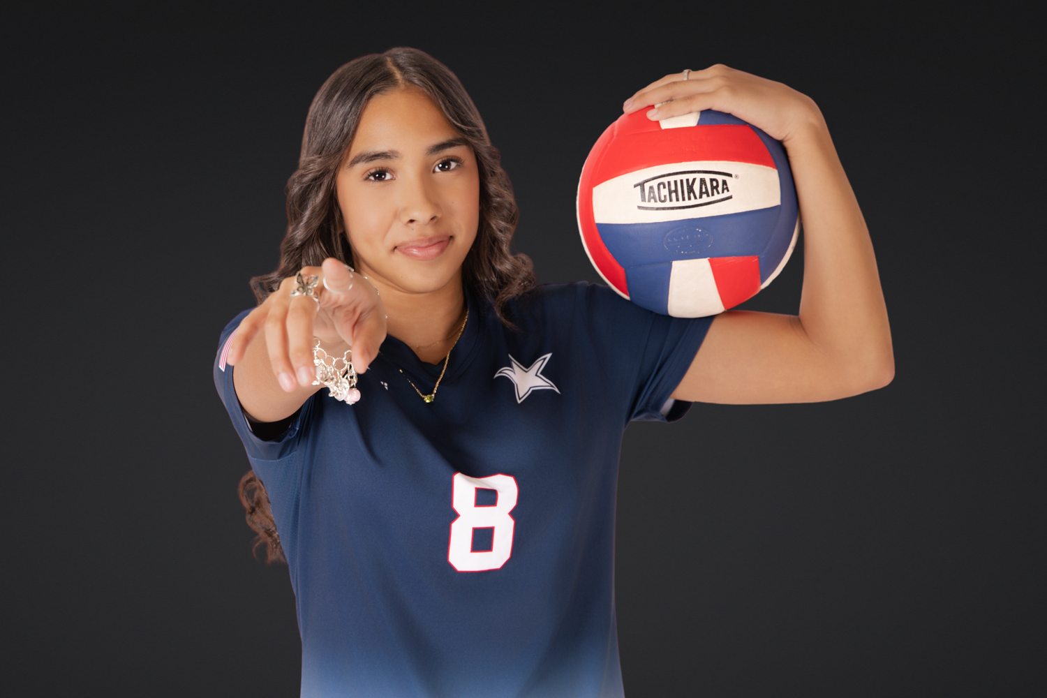 Athlete in navy jersey holds volleyball while pointing at camera against dark background.