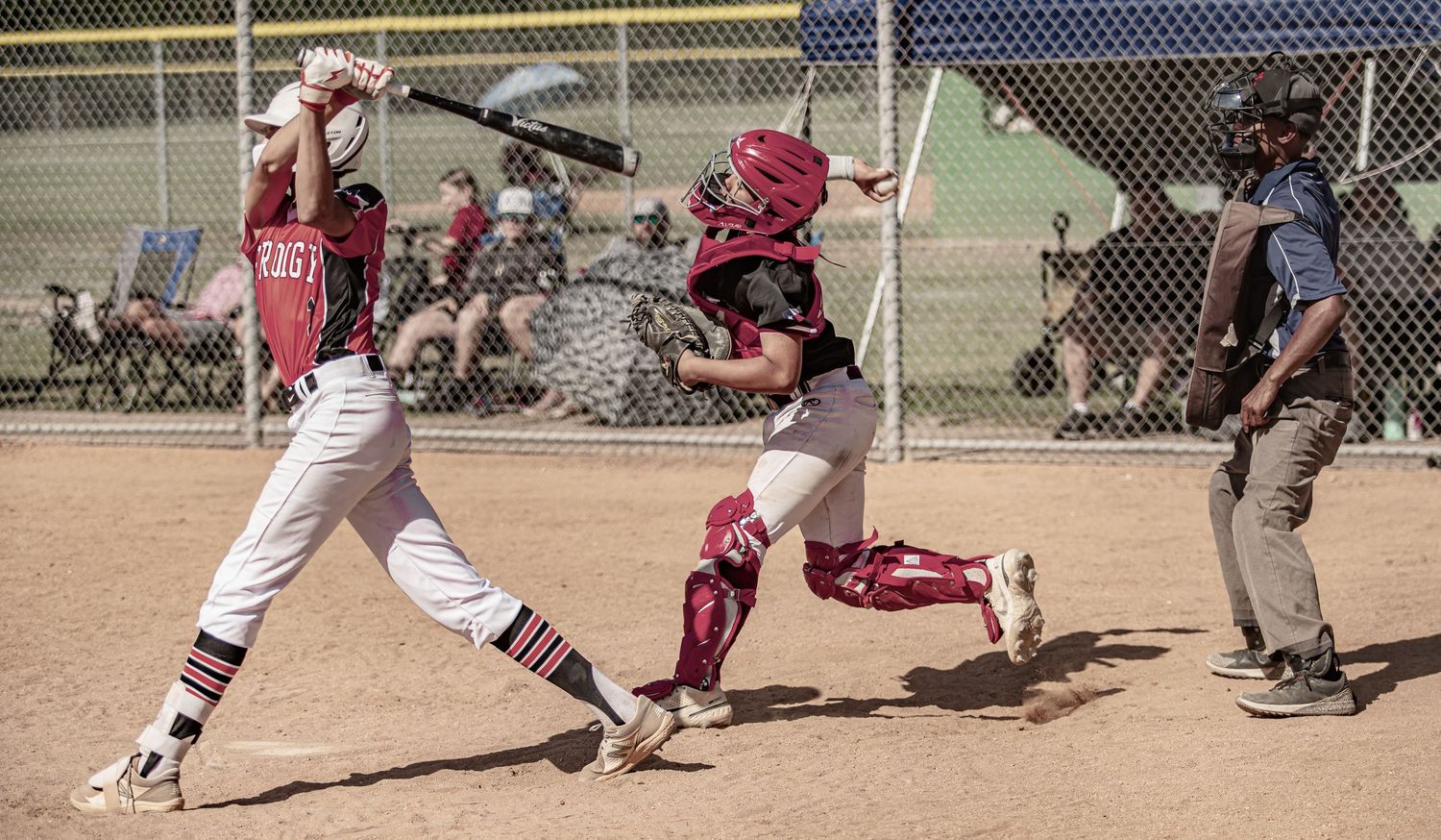 Baseball catcher in red gear reaches to tag batter at home plate during youth baseball game.