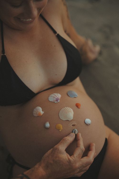 A smiling pregnant woman in a bikini at the beach, with her partner's hand placing a seashell on her bump during an Olive June Moments photo