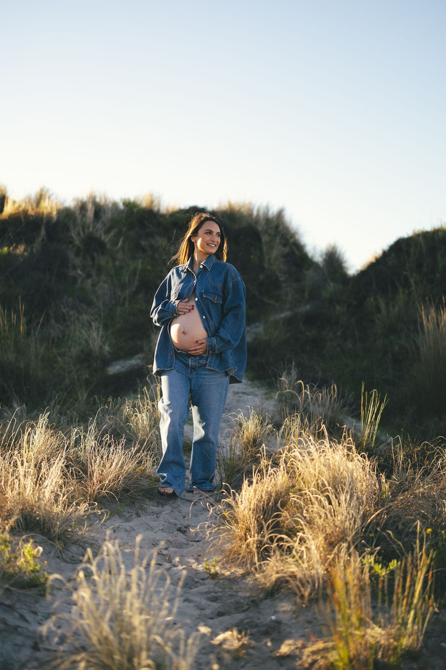 Pregnant woman smiling on a golden hour beach path during an outdoor maternity photo shoot.