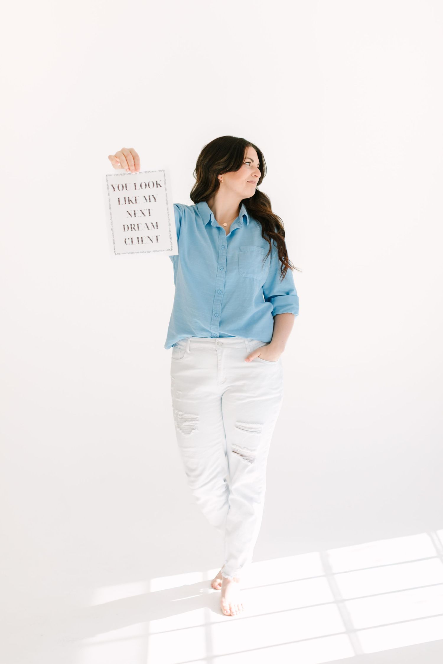 Woman in chambray shirt and white pants poses against white wall with minimal decor and framed artwork.