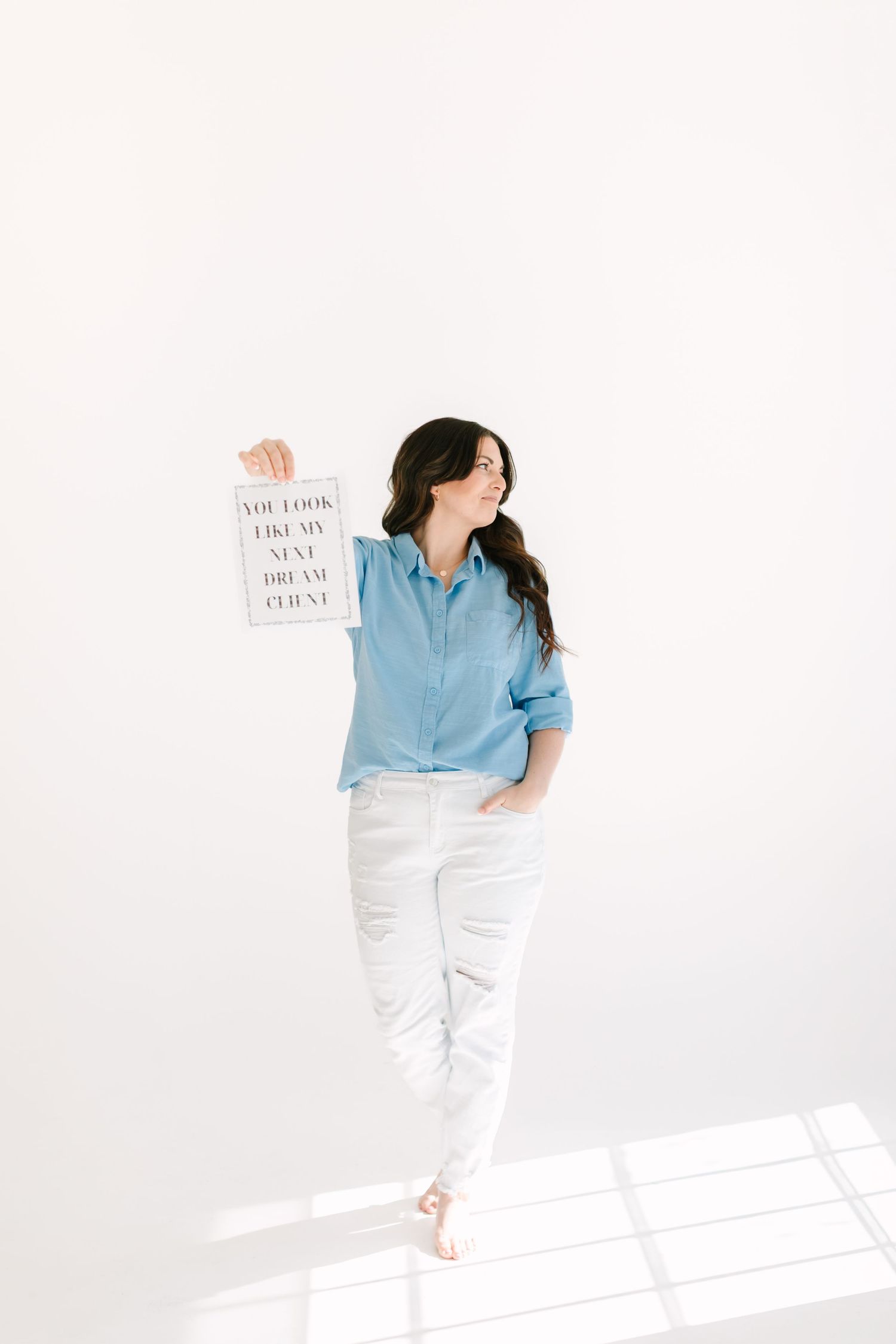 Person in blue shirt and white pants holds up text sign against minimalist white wall.