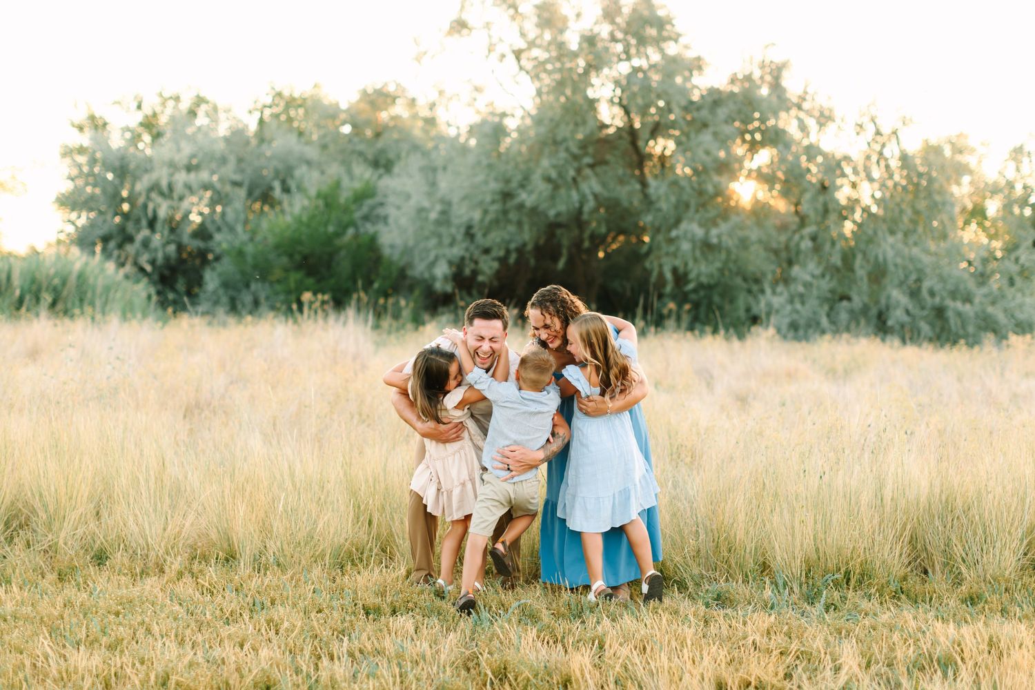 Family laughing together in golden field at sunset, parents and two daughters hugging in warm outdoor portrait in Bountiful, Utah.