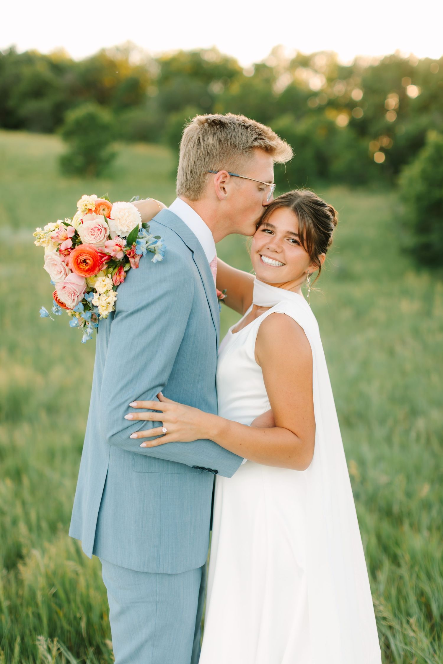 Bride smiling as groom kisses her cheek in a sunlit field, she holds a vibrant bouquet of pink and orange flowers.