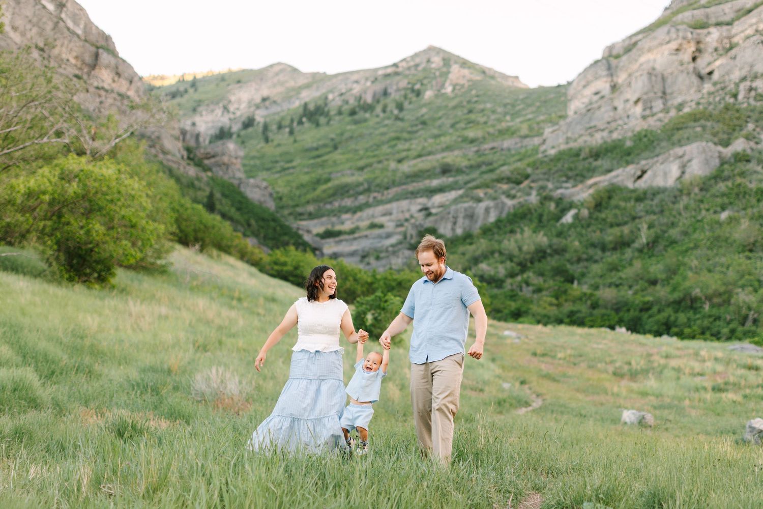 Family of three walking hand-in-hand through green mountain meadow, parents swinging child between them.