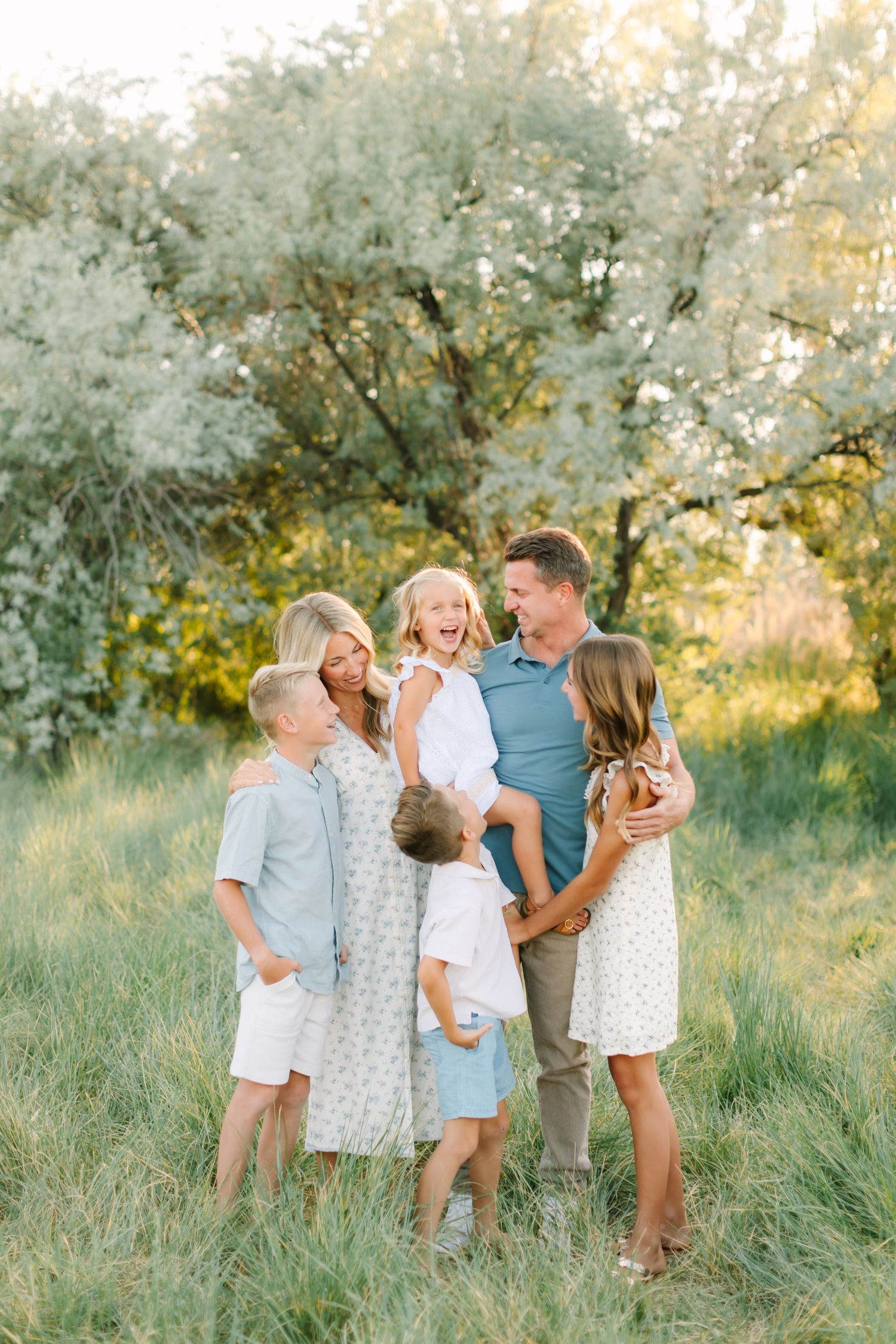 A joyful family of five laughs together in a sunlit meadow with blooming trees, dressed in soft blues and whites.