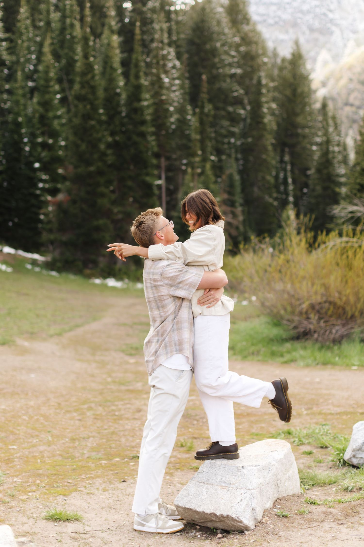 Couple embracing on a rock in a mountain forest setting, wearing white outfits, sharing a joyful moment outdoors.