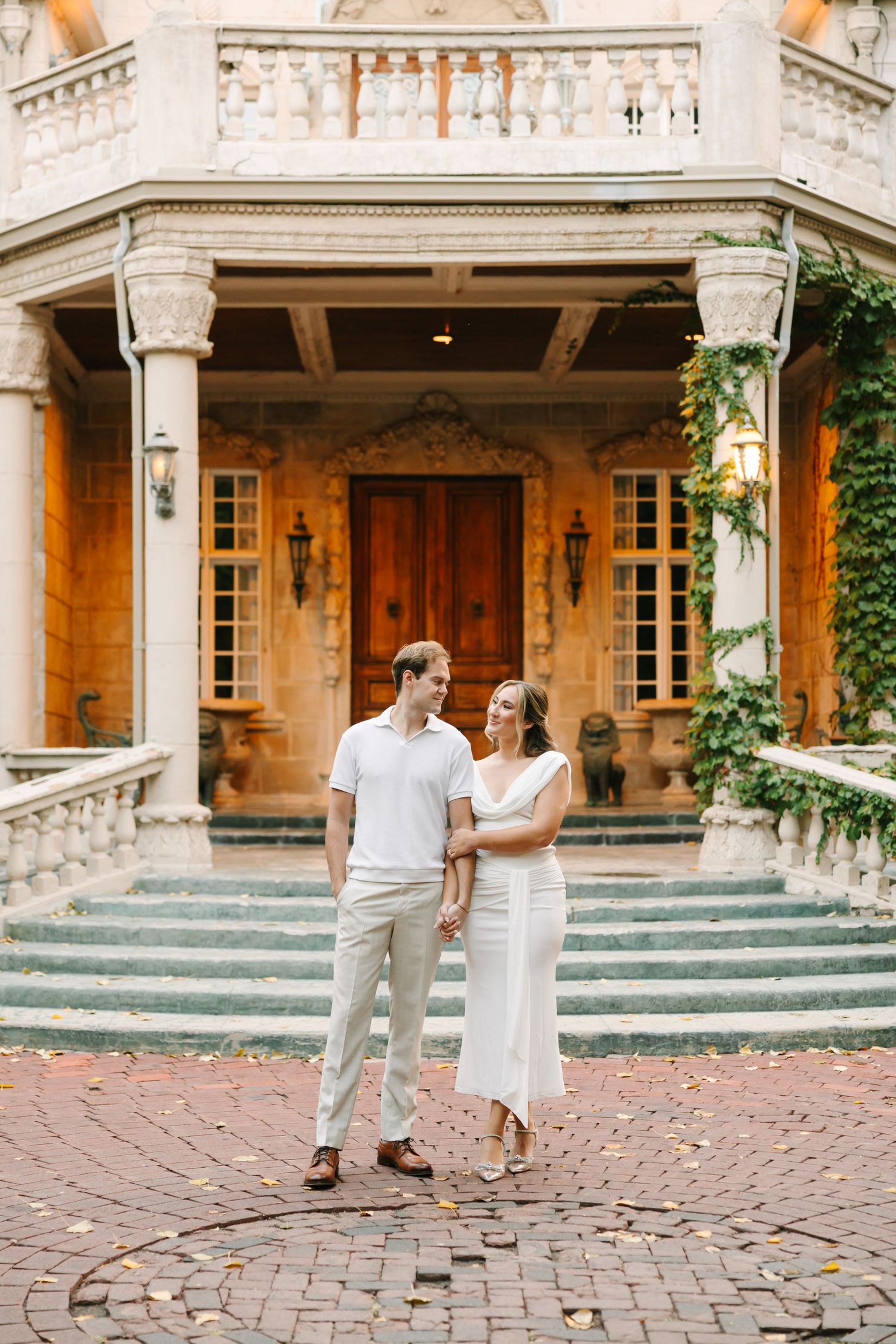 Couple in white outfits dancing and twirling on a cobblestone courtyard in front of a historic stone building.