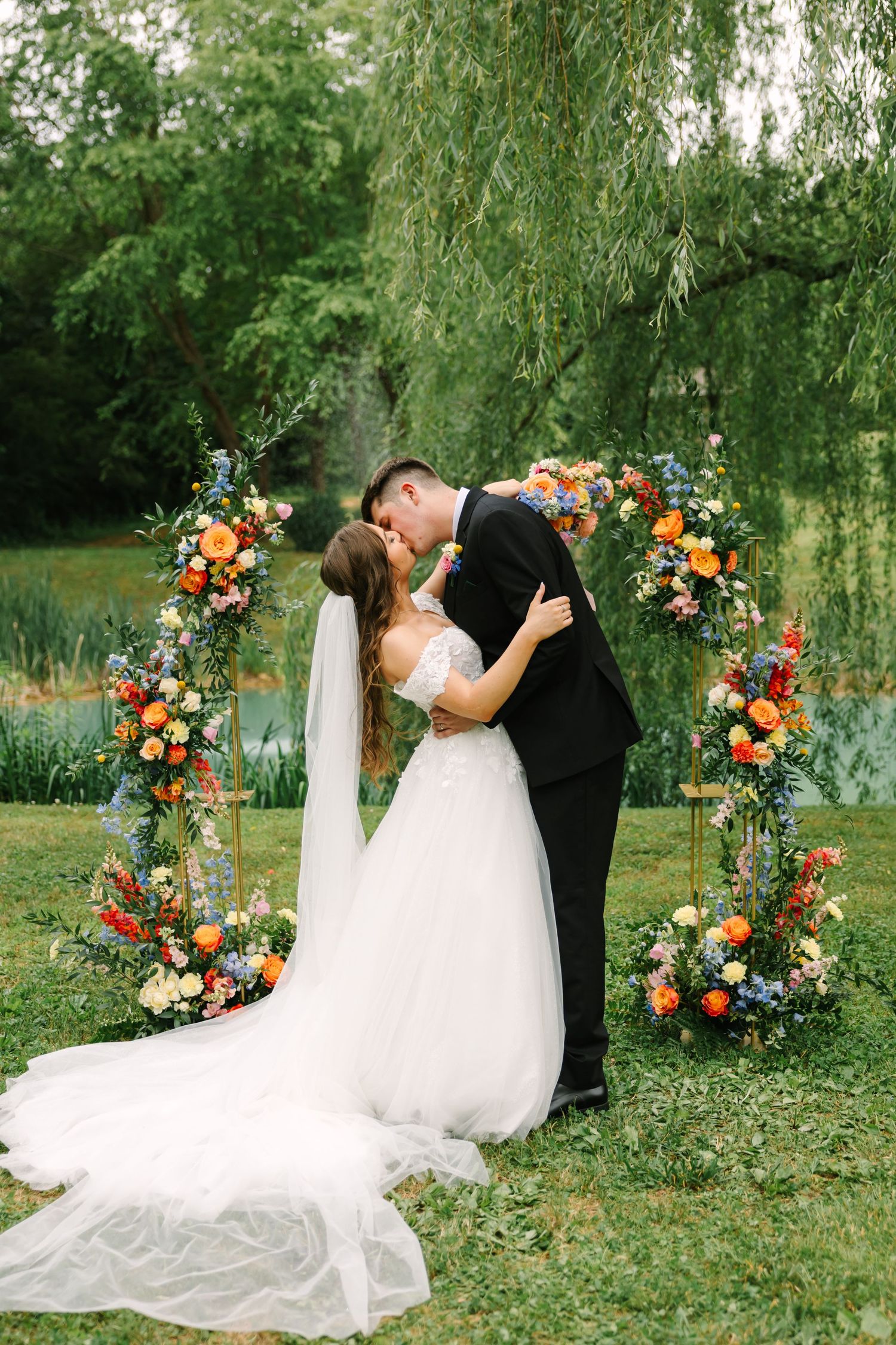 A couple shares a romantic kiss under a floral arch decorated with orange and pink blooms beside a willow tree.