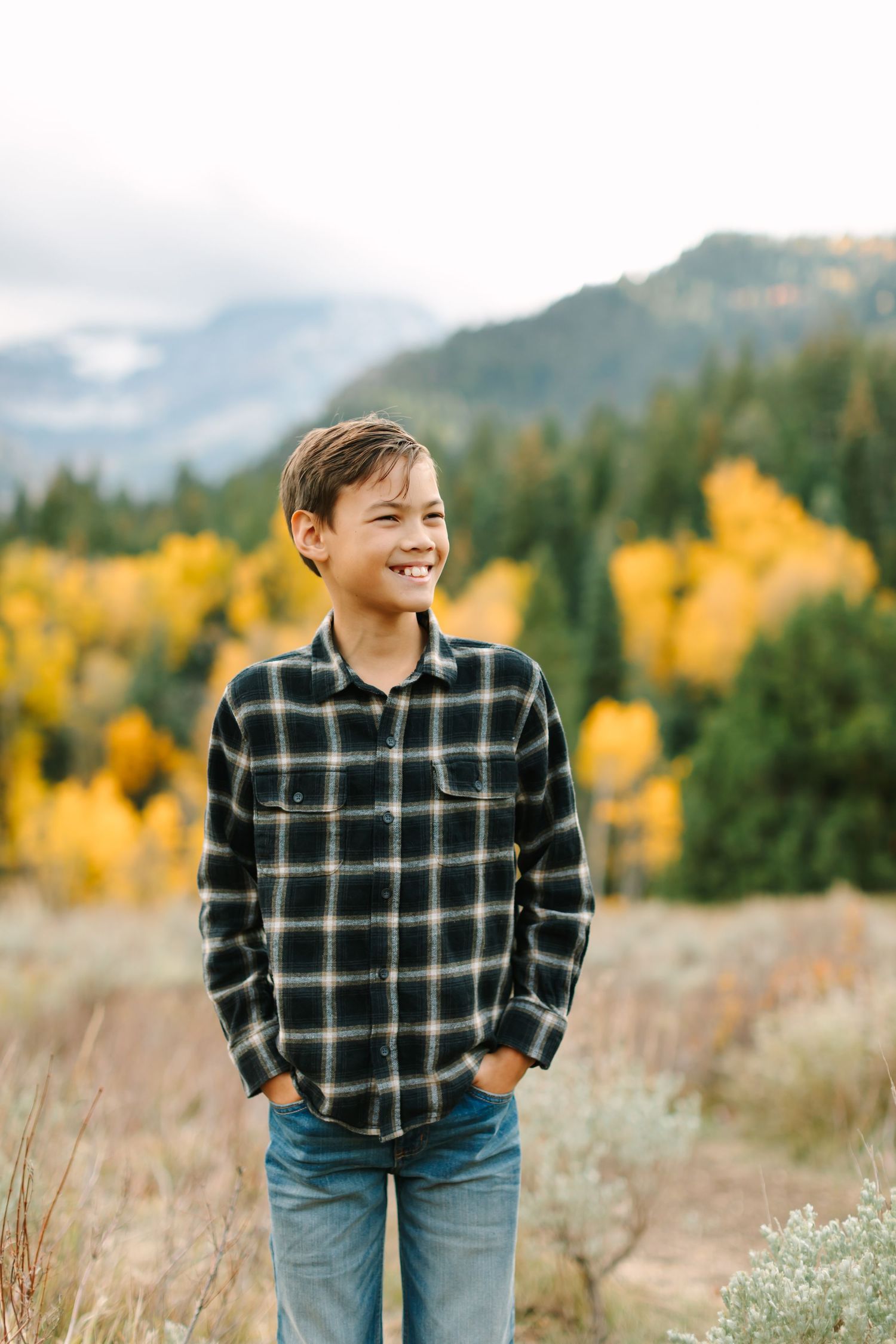 A person in a plaid shirt and jeans stands in a field with autumn foliage and mountains in the background.