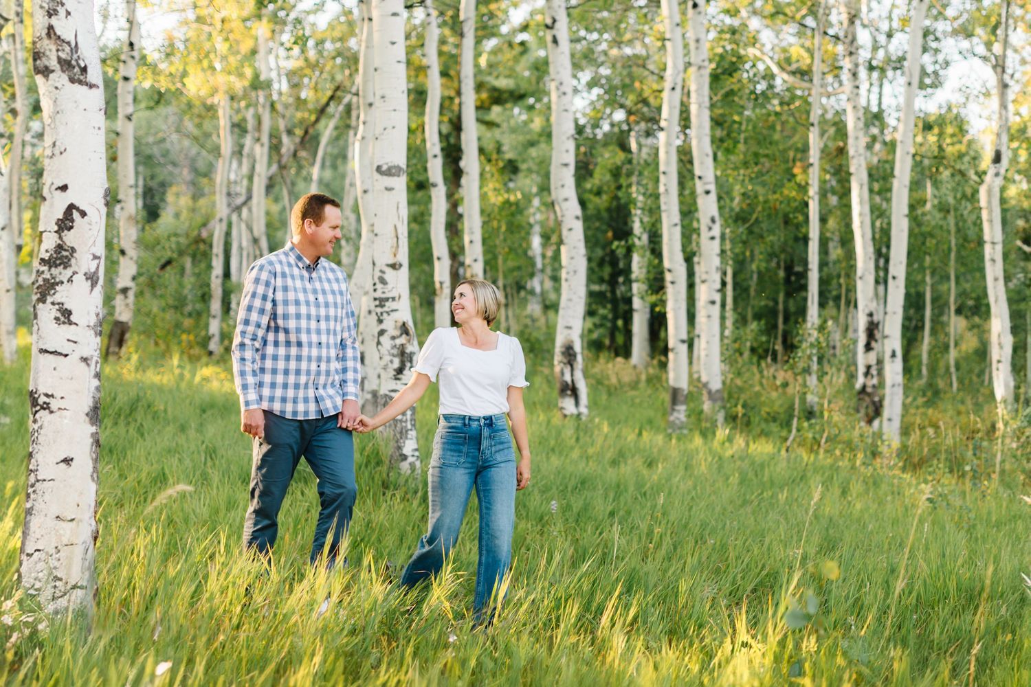Two people hold hands while walking through a serene aspen grove with tall white birch trees on a sunny day.