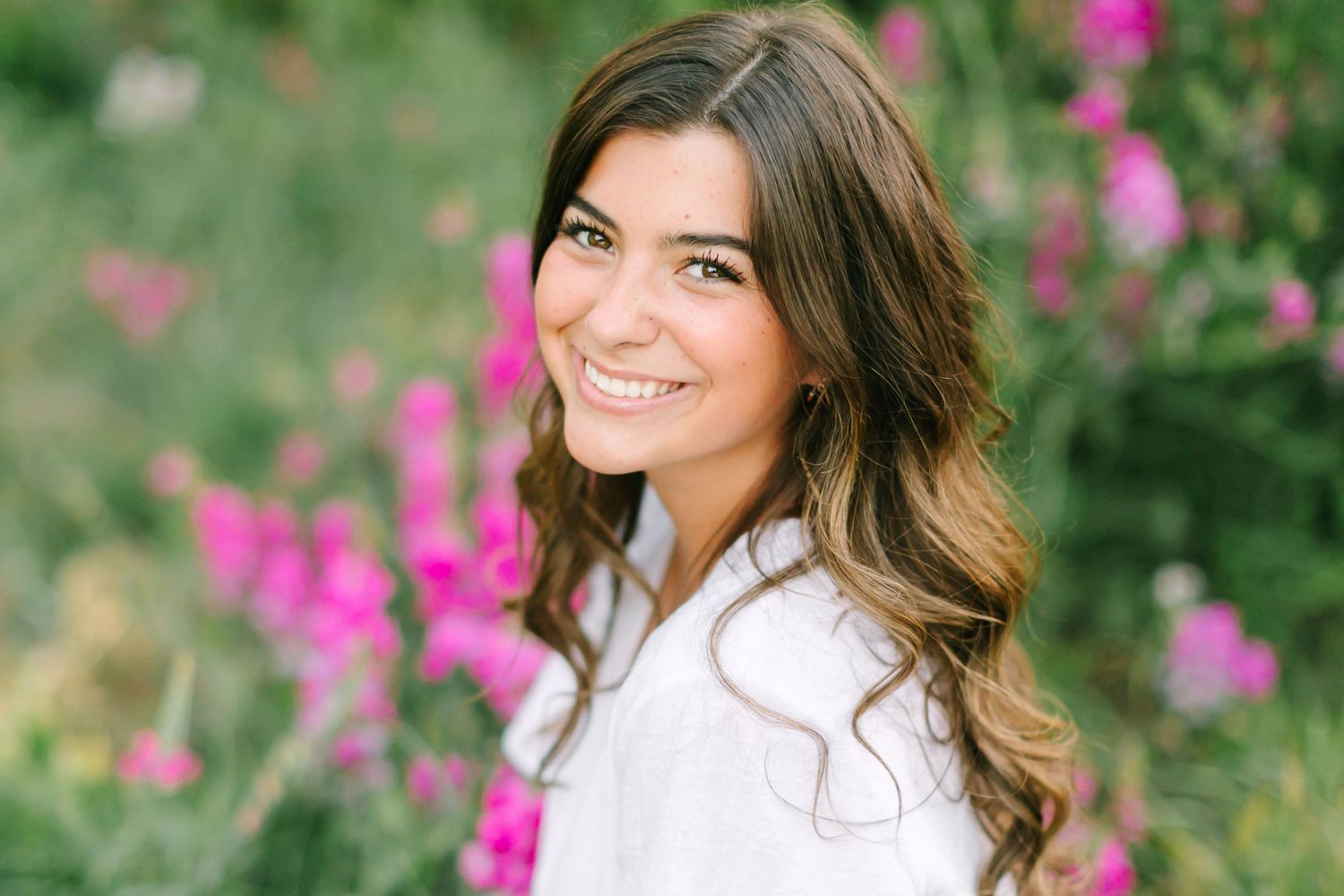A bright outdoor portrait with vibrant pink flowers and greenery creating a soft, natural backdrop.