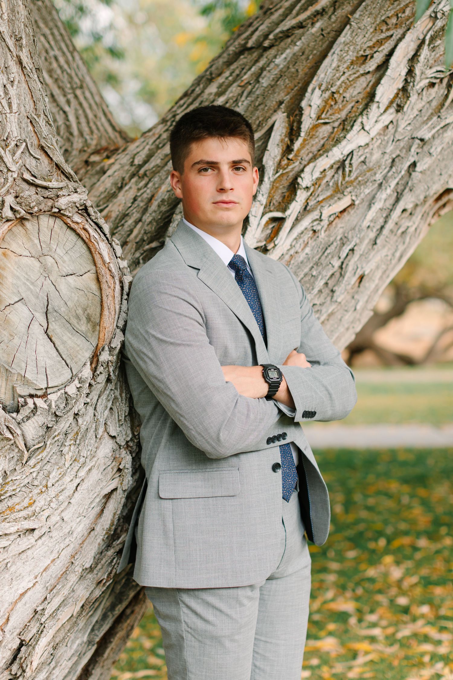 Portrait of a person in a gray suit leaning against a large textured tree trunk with autumn leaves on the ground.
