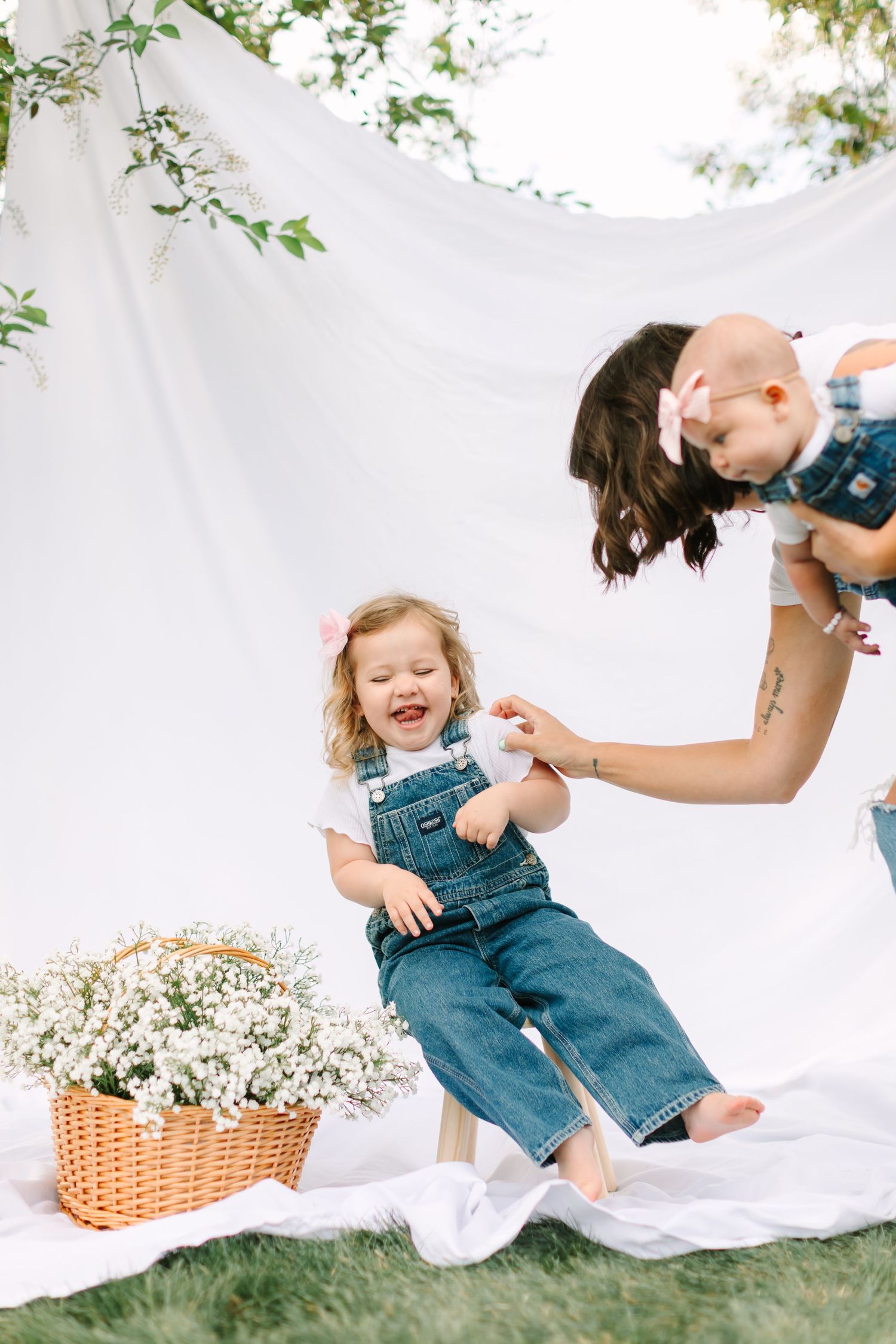 A mother in denim overalls laughs with her two young children outdoors against a white backdrop with flowers.