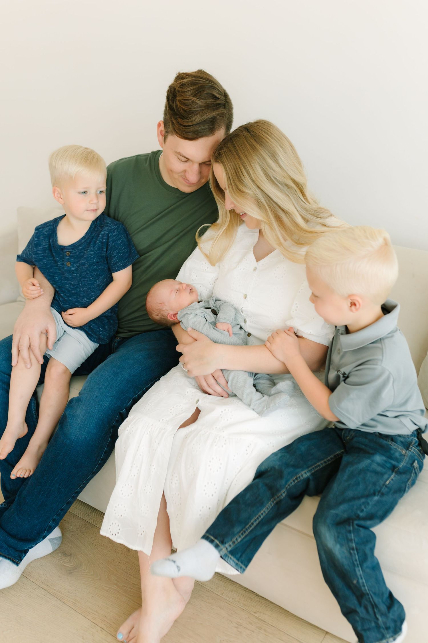 A happy family of four sits together on a couch, parents gazing lovingly at their newborn baby.