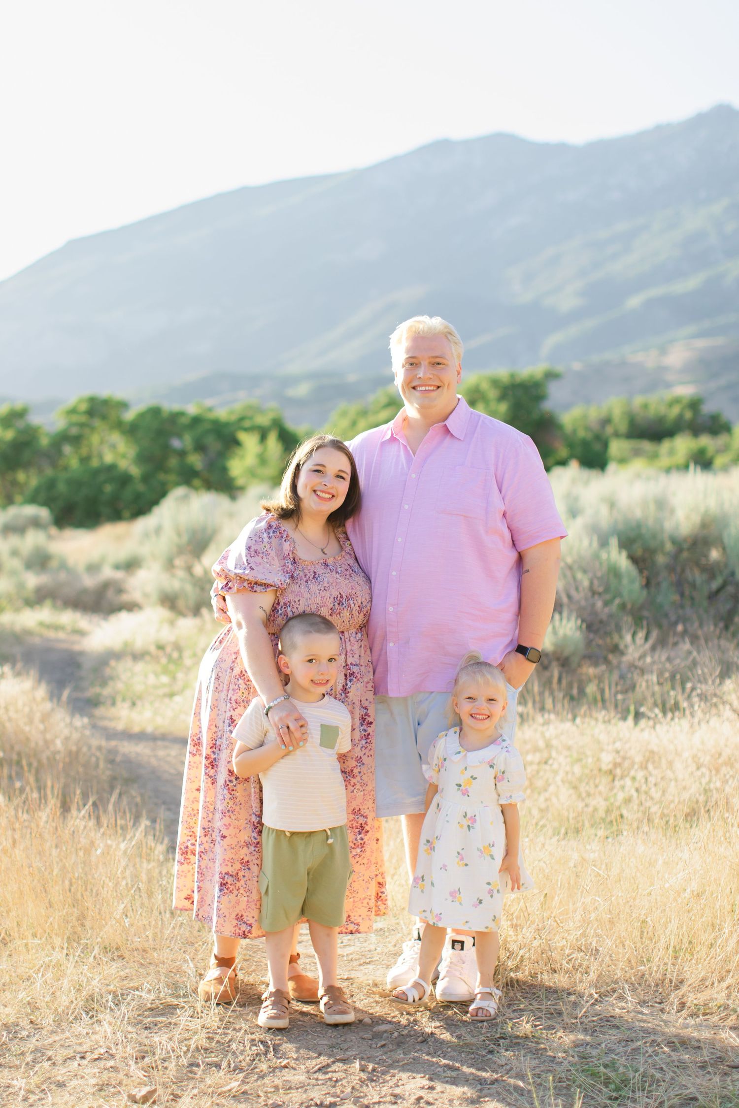 Family stands together in a golden field at sunset with mountains in the background wearing coordinated pink outfits.