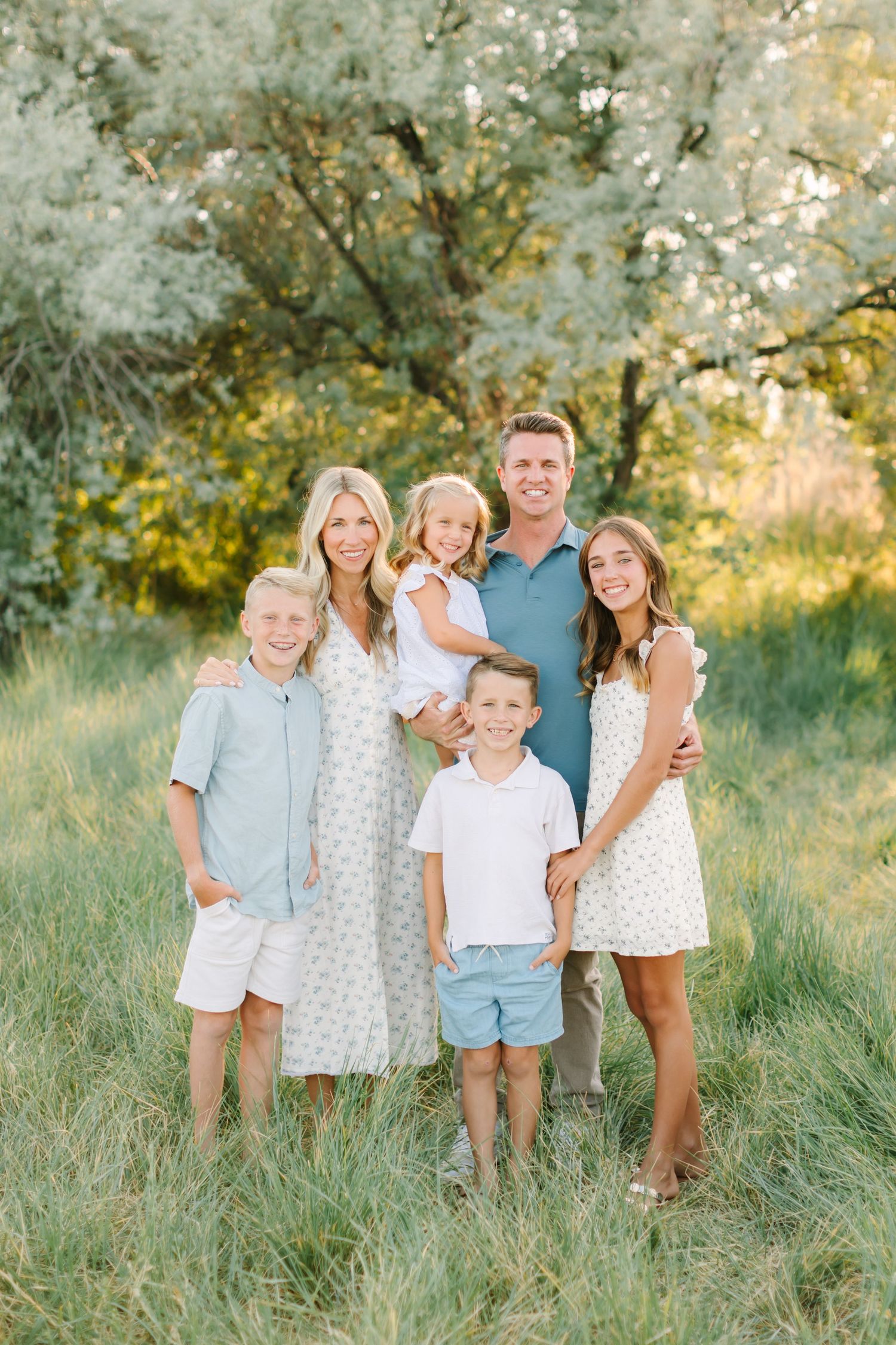 Happy family of six posing outdoors in a green meadow, wearing coordinated light summer outfits.