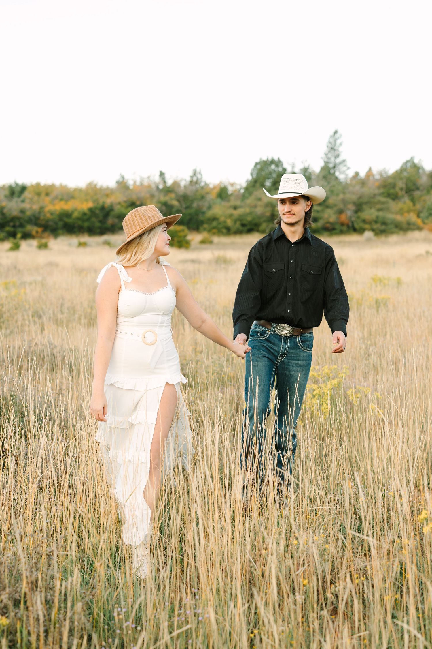 A couple in western attire walks through a golden wheat field during sunset, creating a romantic countryside scene.