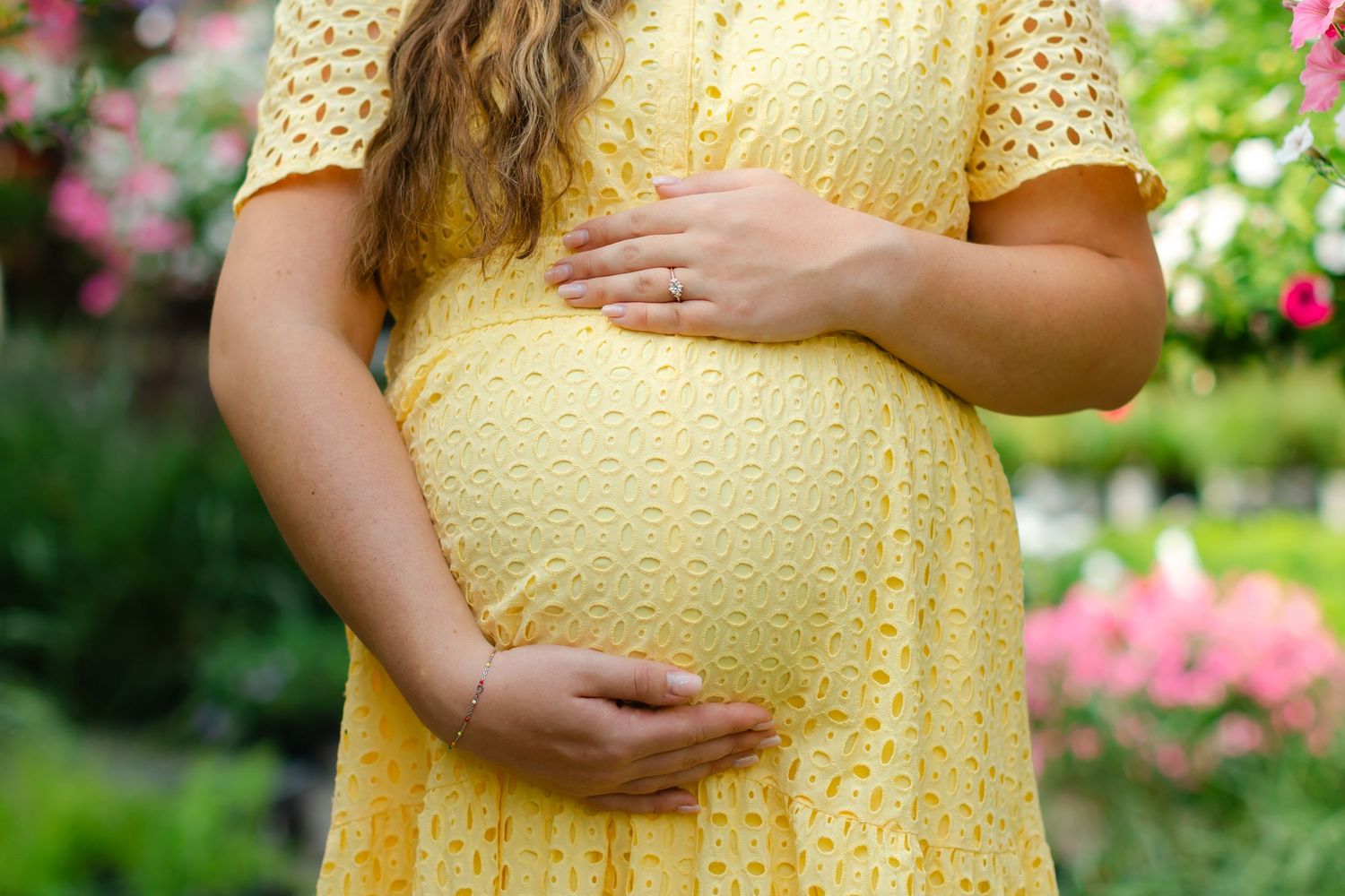 A pregnant woman in a yellow lace dress poses in a garden with pink flowers in the background.