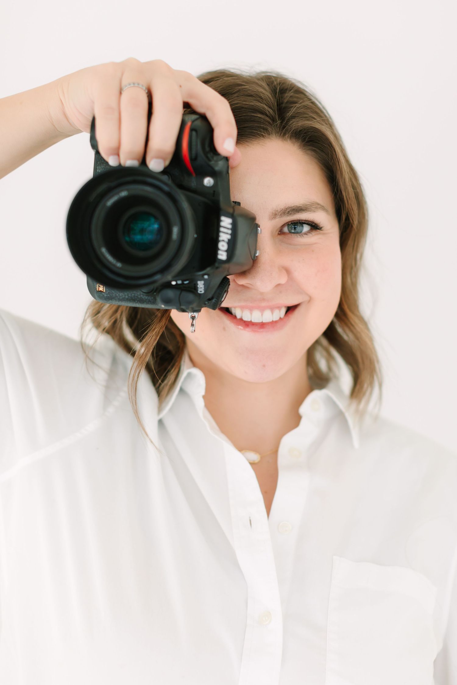 Professional photographer wearing white shirt holds black DSLR camera while taking photos against white background.