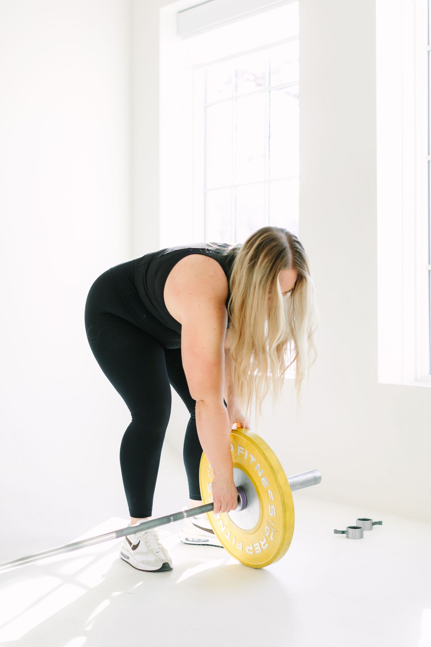 Person in black workout attire loading a yellow weight plate onto a barbell in a bright white gym space.