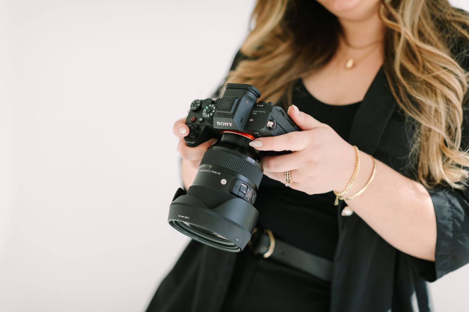 Professional photographer wearing black holds a DSLR camera with mounted lens against white background.