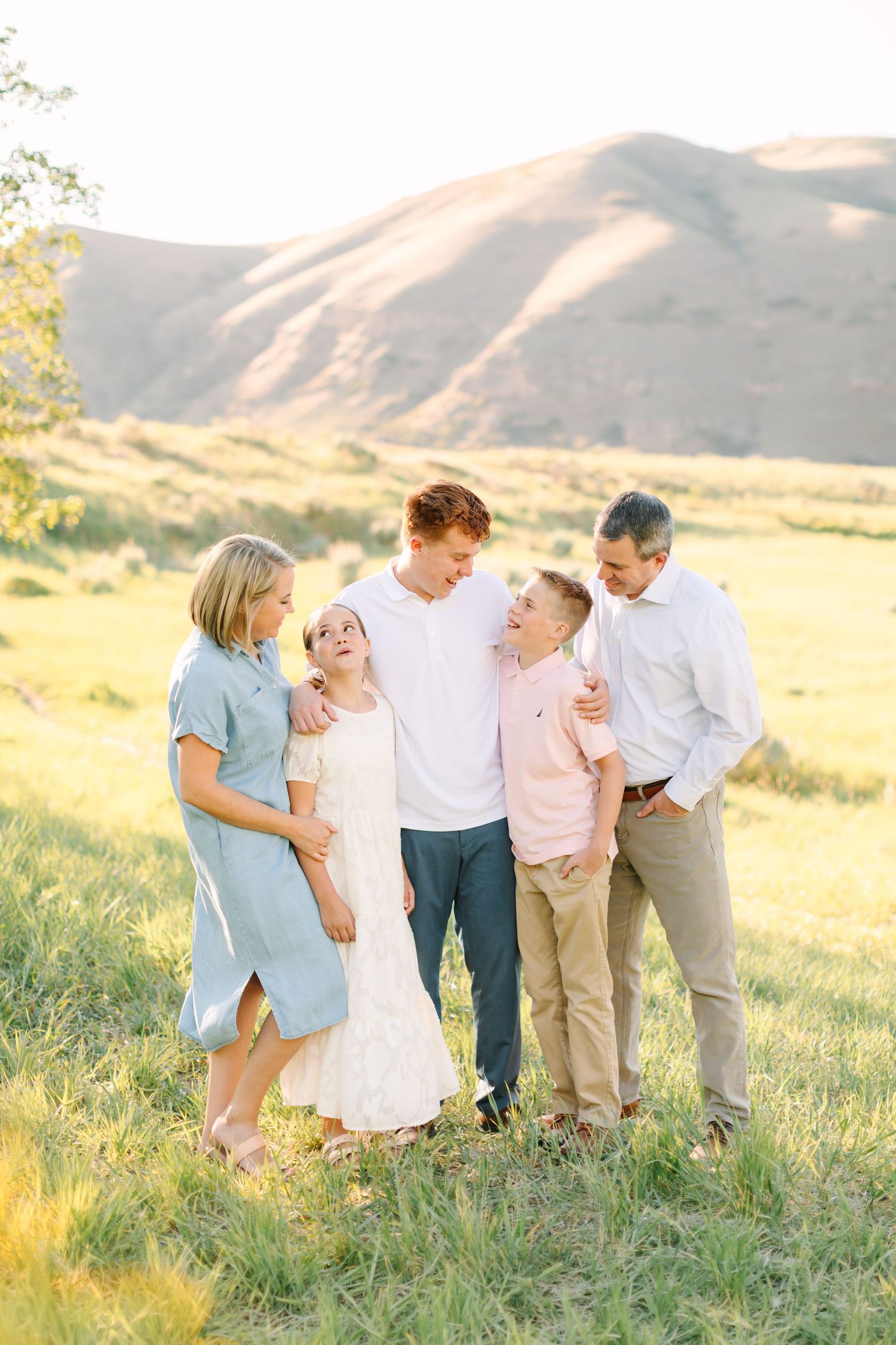 Family of five laughing together in a sunny green meadow with mountain backdrop during golden hour outdoor photo session.