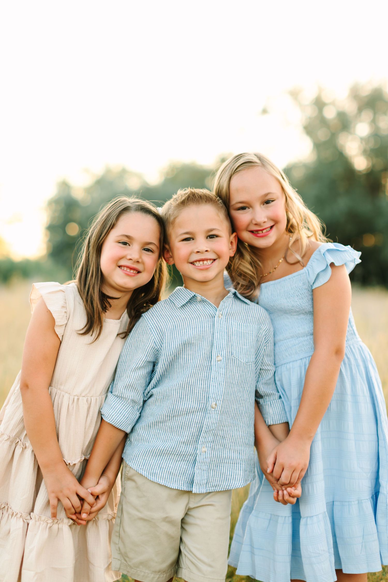 Three smiling children pose together in a sunny field, two girls in blue and cream dresses hugging a boy in a blue shirt in Bountiful, Utah
